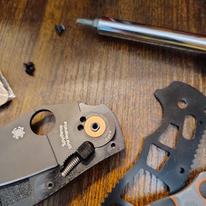May include: Disassembled black folding knife with a partially visible blade, a silver screwdriver, and small screws on a wooden surface. The knife has a circular pivot and the text "Spyderco CPM MagnaCut".