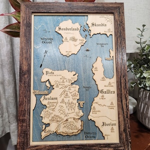May include: A wooden framed map of a fictional land with blue water and brown land masses. The map is labeled with names of places like Skandia, Sonderland, Hibernia, Araluen, Gallica, and Iberion. The map is surrounded by a dark brown wooden frame.