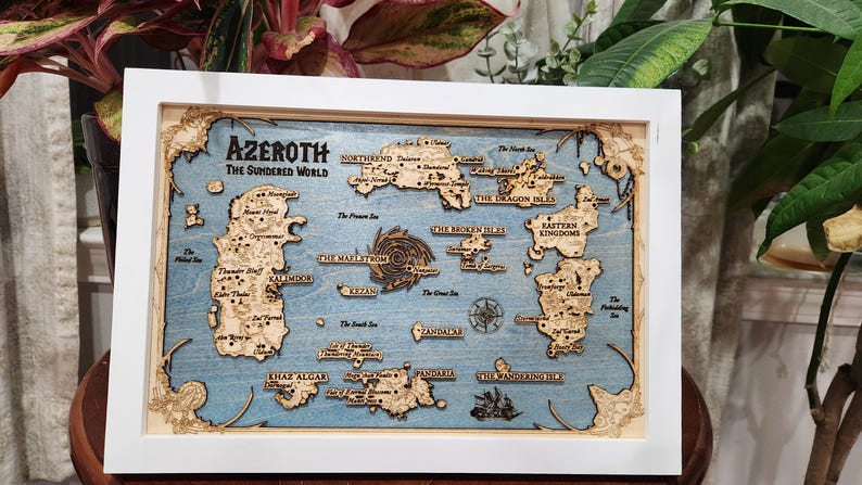 May include: A wooden map of the fictional world of Azeroth from the Warcraft video game series. The map is framed in white and features a blue background with brown islands and text.