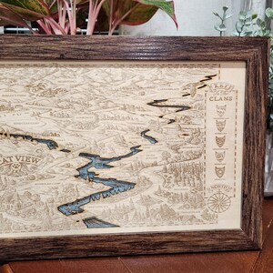 May include: A framed wooden map of the fictional territory of the Warriors Clans, featuring a detailed map of the territory with a river running through it. The map is labeled "Cat View" and includes a compass and a list of the Clans.