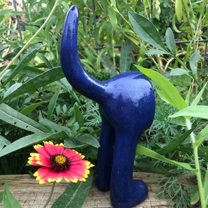Könnte beinhalten: Eine blaue Keramik-Hundefigur mit glitzerndem Finish. Der Hund steht auf seinen Hinterbeinen mit dem Schwanz in der Luft. Die Figur befindet sich in einer Gartenumgebung mit grünen Pflanzen.
