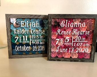 May include: Two framed shadow boxes with paper flowers in blue and pink shades. The boxes display birth announcements for Elijah Kaiden Kearse, born October 26, 2016, weighing 7 pounds 11 ounces and measuring 19.5 inches, and Elianna Renee Kearse, born June 13, 2020, weighing 7 pounds 5 ounces and measuring 20 inches.