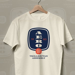 May include: A white t-shirt with a blue square graphic featuring the letters "AERO" and the numbers "19 83". The graphic also includes the text "TROLLHÄTTAN ASSEMBLY" in smaller font.