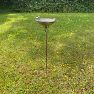 May include: A metal bird bath with two small bird figurines perched on the rim. The bath is mounted on a tall metal pole and is set against a backdrop of green grass.