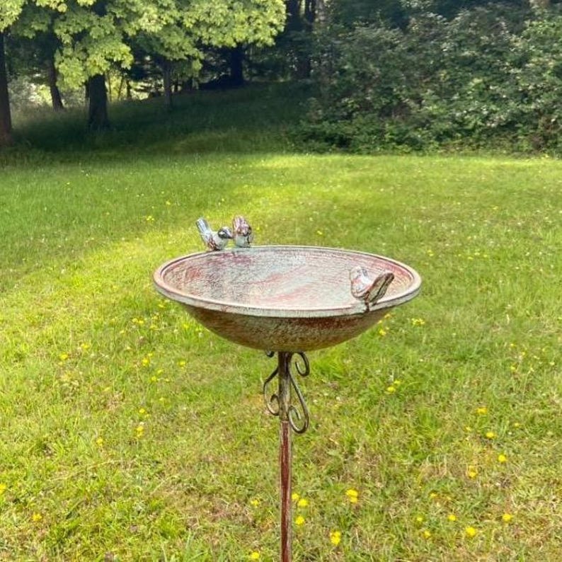 May include: A rustic metal bird bath with a distressed green and red finish. The bath has a round bowl with a raised rim and a decorative metal stand. Three small birds are perched on the rim of the bowl.