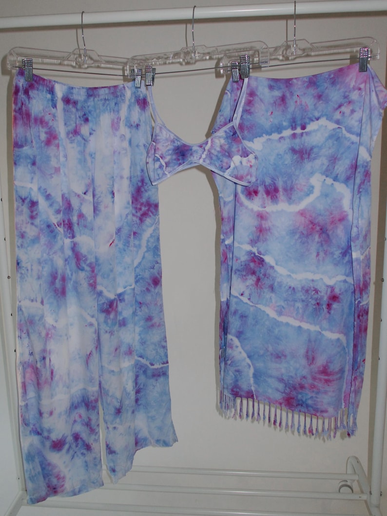 May include: A tie-dye set of clothing with a skirt, pants, and a top. The clothing is a light blue color with pink and white tie-dye patterns. The skirt has a fringe trim.