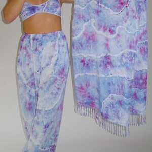 May include: A tie-dye set with a matching top, pants, and scarf. The set is in shades of blue and purple with a white background. The scarf has a fringe trim.