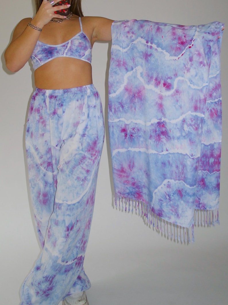 May include: A tie-dye set with a matching top, pants, and scarf. The set is in shades of blue and purple with a white background. The scarf has a fringe trim.