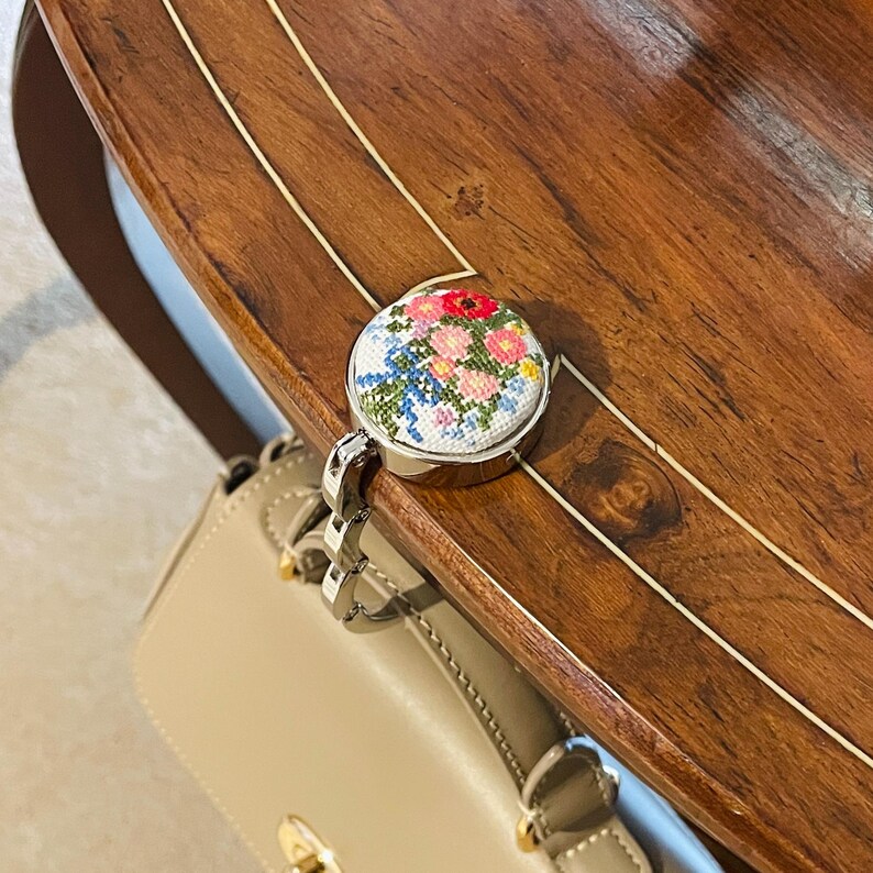 May include: A silver handbag hook with a round floral design. The hook is attached to a beige handbag.