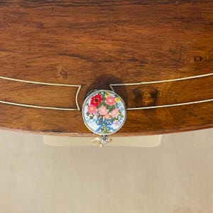 May include: A round, silver-colored drawer pull with a floral design. The flowers are pink, red, and blue, and are embroidered on a white background.