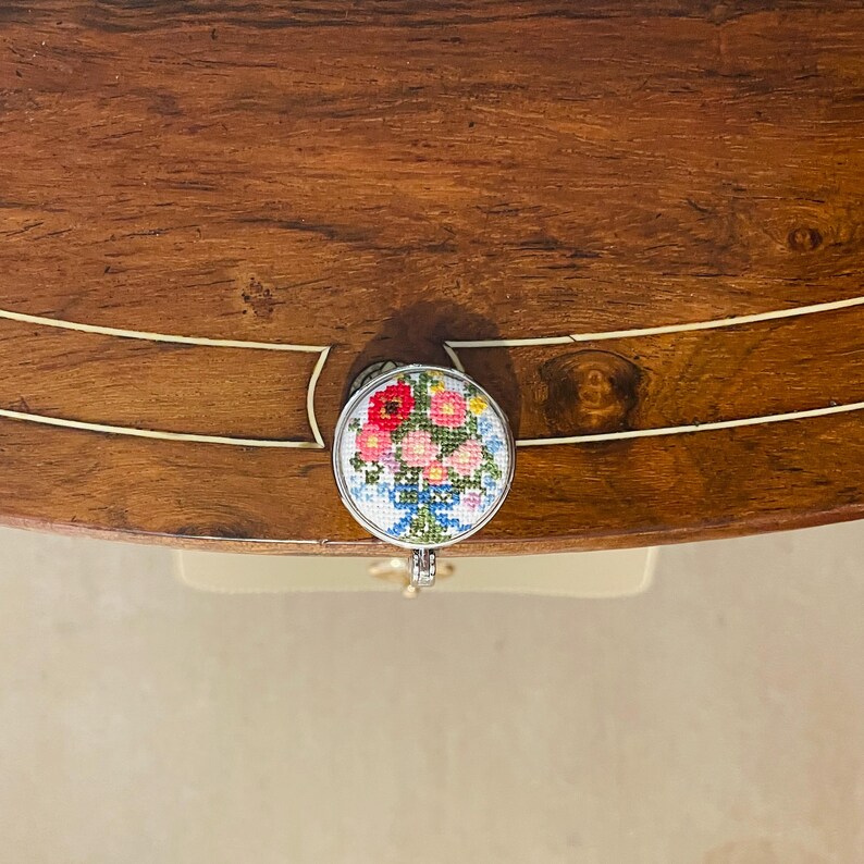 May include: A round, silver-colored drawer pull with a floral design. The flowers are pink, red, and blue, and are embroidered on a white background.
