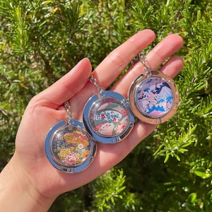 以下が含まれることがあります： 日本のデザインが施された、3つの丸いシルバーカラーのバッグハンガー。それぞれ異なる画像が特徴です。花柄、扇子柄、桜の山並みの風景。ハンガーは、緑の葉を背景に、手で持たれています。