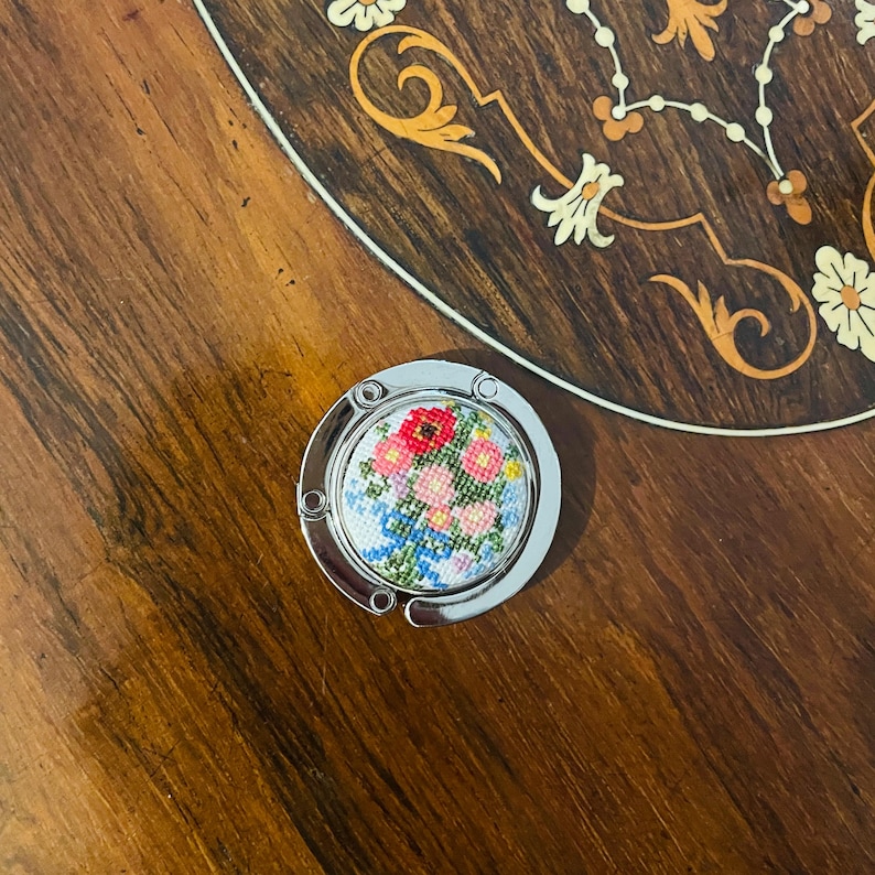 May include: A silver-toned purse hook with a round, white background and a colorful floral embroidery design. The flowers are pink, red, and blue.