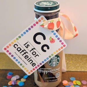May include: A Starbucks Frappuccino chilled coffee drink in a 13.7 fluid ounce glass bottle with a green cap. A white rectangular gift tag with a colorful polka dot border is tied to the bottle with an apple-patterned ribbon. The tag reads Cc is for caffeine and From: Sophia Jameson.