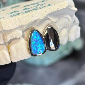 Peut inclure: Deux dents de grill en or, l'une avec une pierre d'opale bleue et l'autre avec une finition argent&eacute;e.