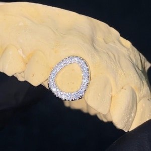 May include: A silver tooth grill with a round, open design and a row of clear diamonds around the edge.