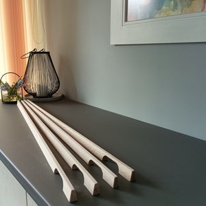 Pode incluir: Cinco puxadores de porta de armário de madeira sem acabamento com um design simples e curvo. Os puxadores estão dispostos em uma fila sobre uma superfície cinza.