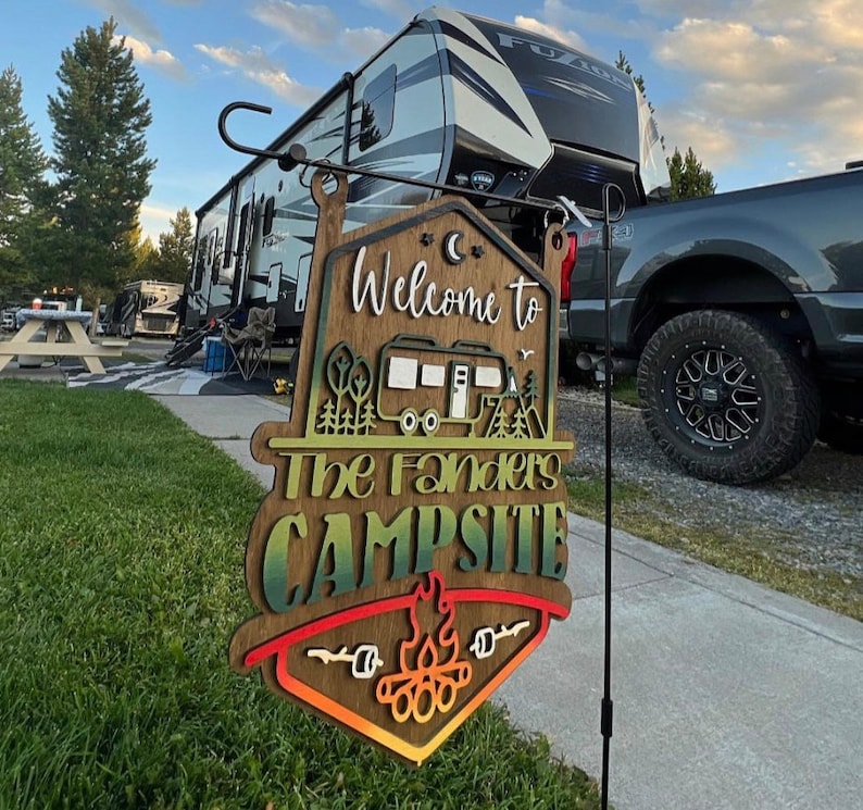 May include: A wooden welcome sign with a rainbow gradient and the text "Welcome to The Fanders Campsite". The sign features a small camper van, trees, and a campfire with marshmallows.