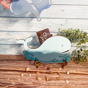 May include: A light blue whale-shaped wooden card holder with a matching water spout design. The whale has a gradient effect and is supported by wooden legs. Small seashells are scattered around the base.