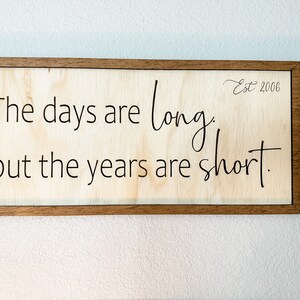 May include: Wooden framed sign with the quote "The days are long, but the years are short." The sign has a light wood background and dark brown frame. The text is in black, with "Est 2006" in the upper right corner.