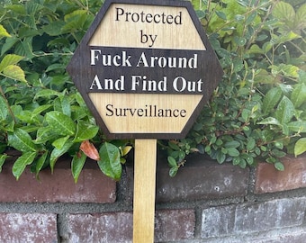 May include: A wooden garden sign with the text "Protected by Fuck Around And Find Out Surveillance". The sign is a hexagon shape with a dark brown border and a light brown centre, mounted on a wooden stake.