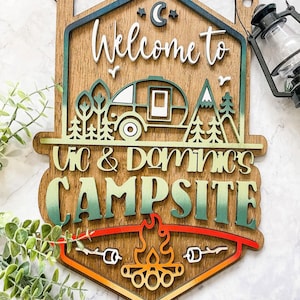 May include: Wooden sign with a camping theme. It says "Welcome to Vic & Dominics Campsite" with a camper, trees, mountains, and a campfire.