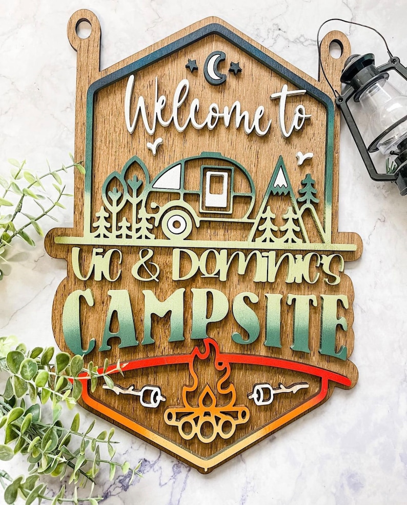 May include: Wooden sign with a camping theme. It says "Welcome to Vic & Dominics Campsite" with a camper, trees, mountains, and a campfire.