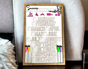 May include: Wooden-framed whiteboard with the text "Success" and "January" through "December" in large letters. The board includes colorful markers and small exercise icons. The text reads "Success doesn't come from what you do occasionally - it comes from what you do consistently."