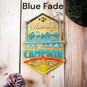 May include: Wooden sign with a blue fade effect, featuring the text "Welcome to The Koha Campsite" with a campfire graphic at the bottom.