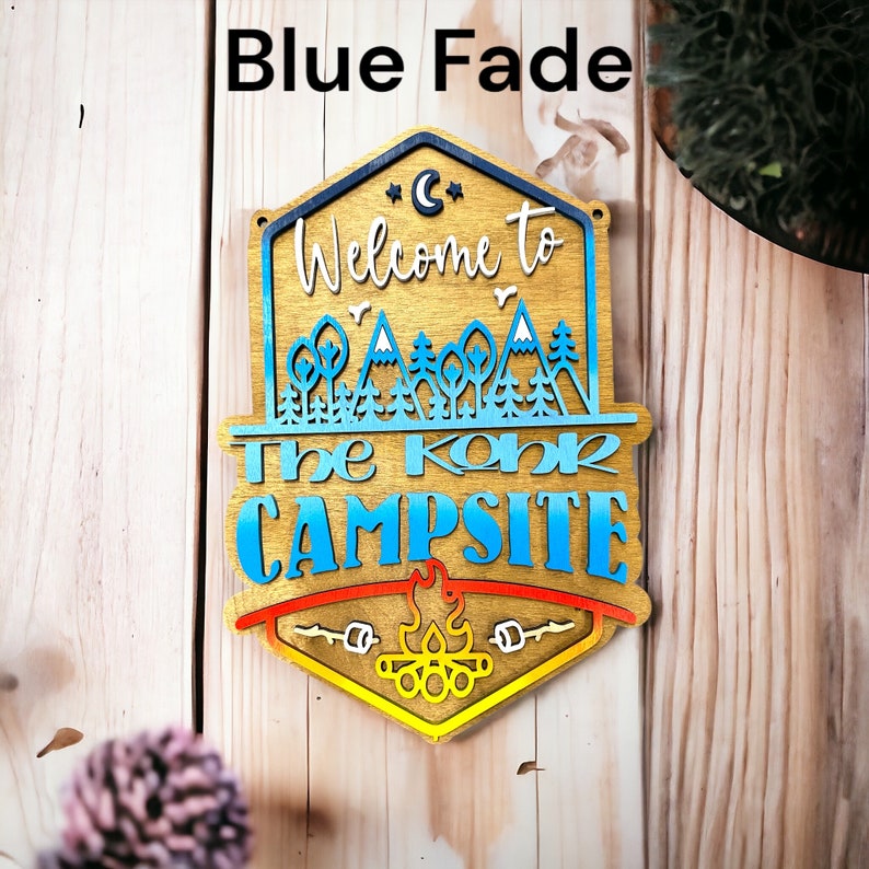 May include: Wooden sign with a blue fade effect, featuring the text "Welcome to The Koha Campsite" with a campfire graphic at the bottom.