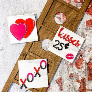 May include: Wooden Valentine's Day decor featuring a brown wooden frame with circular cutouts and three white square signs. The signs display a red and pink heart, the text "Kisses 25¢" with a lip graphic, and the letters "xoxo" in red and pink.