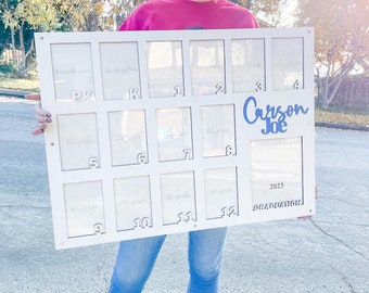 May include: White wooden photo frame with 12 clear picture slots for each grade from Pre-Kindergarten to 12th grade. The frame has the text "Carson Joe 2023 Graduation" in blue lettering.
