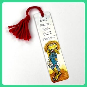 Puede incluir: Un marcapáginas caprichoso con una rana de dibujos animados con un peto sosteniendo una margarita, con el texto "Have I toad you lately that I love you?" Una borla roja está unida a la parte superior del marcapáginas.