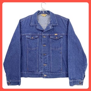 May include: A medium blue denim jacket featuring a classic collar, long sleeves with buttoned cuffs, and a front button closure. The jacket includes two chest pockets with buttoned flaps and a small yellow brand label on the left chest pocket.