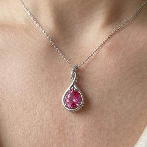 Pode incluir: Um colar de tom prateado com um pendente em forma de lágrima e uma pedra central rosa vibrante. O pendente é emoldurado por um design metálico em espiral com pequenas pedras claras, suspenso por uma delicada corrente prateada.