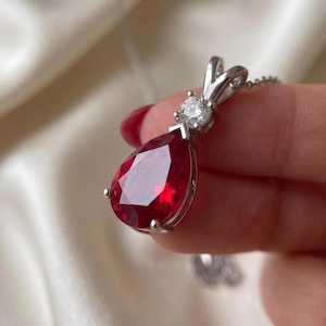 May include: A silver-toned pendant necklace featuring a large, pear-shaped, deep red gemstone as the centrepiece. Above the red stone, a single, small, round, clear white gemstone is set in a silver-toned metal mount attached to a delicate chain.