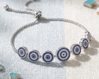 Pode incluir: Uma pulseira ajustável de tom prateado com uma série de berloques circulares. Cada berloque é decorado com anéis concêntricos de pequenas pedras azuis e brancas, criando um padrão circular que lembra um motivo de olho em uma corrente.