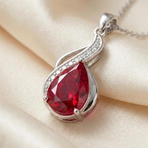 May include: A silver-toned pendant necklace featuring a large, pear-shaped, deep red gemstone set in a metallic frame. The upper portion of the pendant is curved and accented with small, clear, sparkling stones, attached to a delicate silver chain.