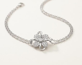 May include: A silver-toned bracelet featuring a central four-petal flower charm encrusted with small clear stones. The bracelet is constructed with a delicate double-chain design that connects to the flower centerpiece, finished with a clasp.