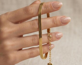 May include: A gold-toned herringbone chain bracelet featuring a flat, woven link design. The bracelet includes an adjustable extender chain with a small, solid heart-shaped charm at the end and a circular spring ring clasp mechanism.