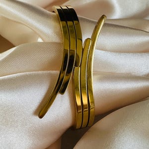Könnte beinhalten: Ein goldfarbenes Metall-Armreif-Armband mit einem einzigartigen, geschwungenen Design. Das Armband besteht aus drei separaten Bändern, die miteinander verschlungen sind.