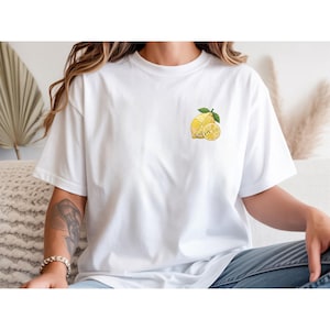 Puede incluir: Camiseta blanca con un pequeño diseño de limón bordado en el pecho. La ilustración del limón incluye un limón entero, un limón en rodajas y hojas verdes. Una prenda casual para el día a día.