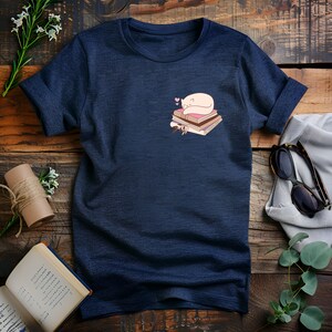 May include: Navy blue t-shirt featuring a small graphic of a sleeping white cat on a stack of books with pink hearts. The t-shirt has short sleeves and a crew neck. The design is on the left chest area.