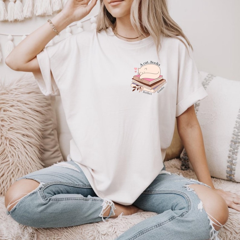 May include: Cream-colored t-shirt featuring a graphic of a sleeping cat atop a stack of books. The text "A cat, Books, Perfect Combo" is printed on the shirt. The shirt is paired with blue jeans.