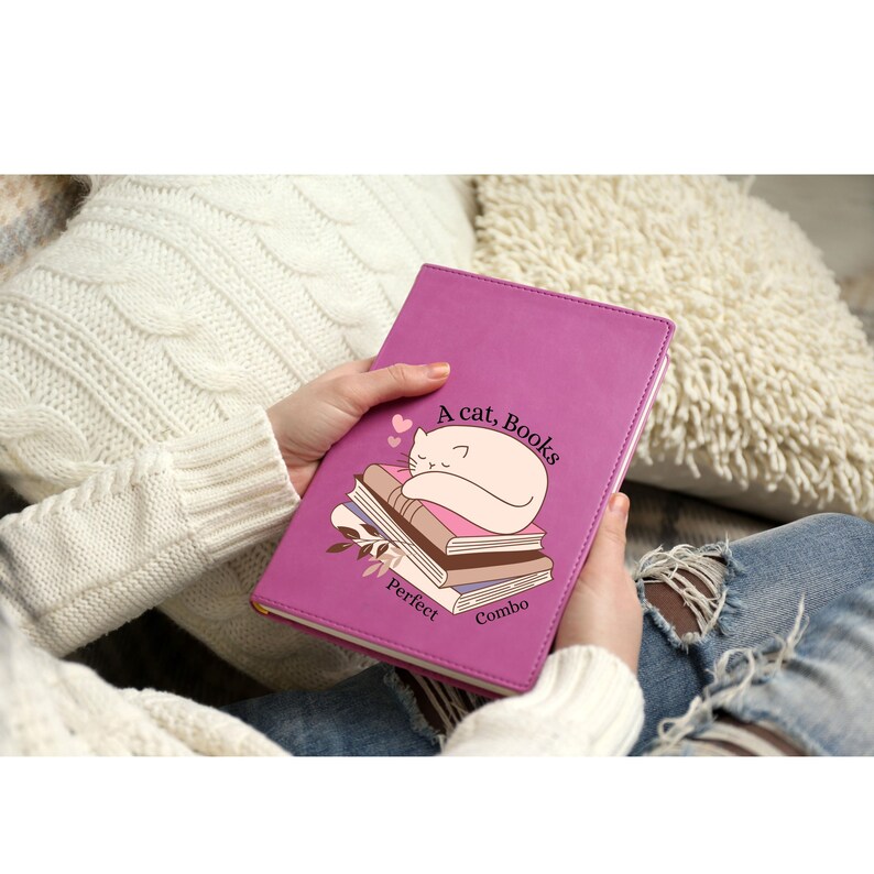 May include: A magenta-colored journal with the words "A cat, Books, Perfect Combo" and an illustration of a sleeping cat on a stack of books. The journal is held in someone's hands.