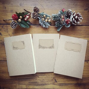 Peut inclure: Trois carnets beiges avec des étiquettes décoratives sont disposés sur une surface en bois. Au-dessus, des brins festifs avec des pommes de pin, des baies et des accents dorés ajoutent une touche saisonnière. Les carnets sont vierges.