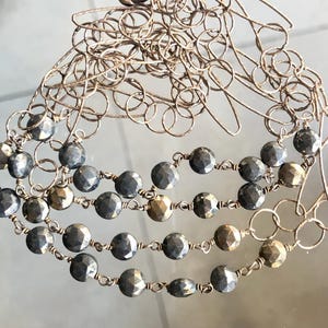 Puede incluir: Un collar de cadena de plata con múltiples hileras de cuentas de pirita facetadas. Las cuentas son de color gris oscuro y tienen un brillo metálico.