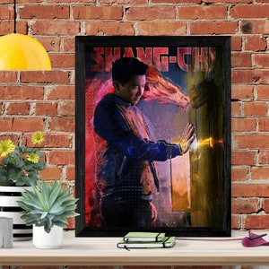 以下が含まれることがあります： 青いジャケットを着た男性が「SHANG-CHI」という赤い文字と白い文字で書かれたポスターが額装されています。男性は、オレンジと黄色の光る背景に、後ろに龍の頭が描かれた背景に立っています。