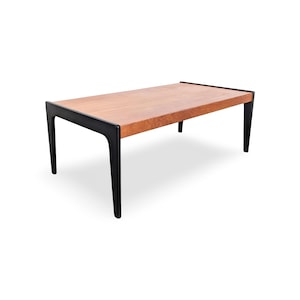 Peut inclure: Une table basse rectangulaire dotée d'un plateau en bois brun clair naturel et d'un cadre noir contrastant. Le cadre comprend quatre pieds fuselés qui s'étendent vers le haut pour former les bords latéraux de la table, soutenant le plateau.