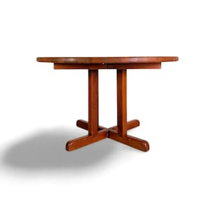 Könnte beinhalten: Ein runder Esstisch aus Holz mit warmem, rötlich-braunem Finish. Der Tisch hat eine zweiteilige Platte und ein Säulenfuß mit vier Beinen. Der Tisch hat einen Durchmesser von ca. 102 cm.